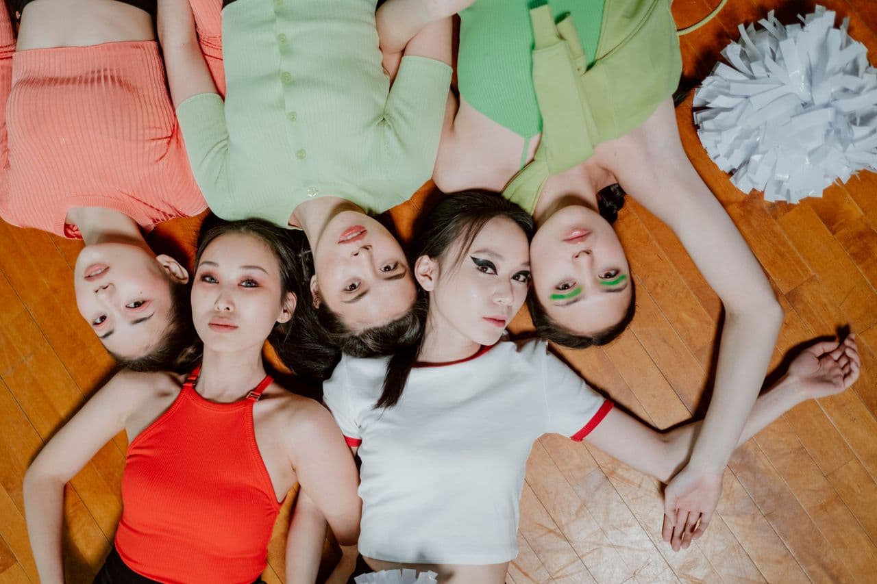 Five people in coordinated pastel outfits lying in a circle for a K-pop-style group photo.