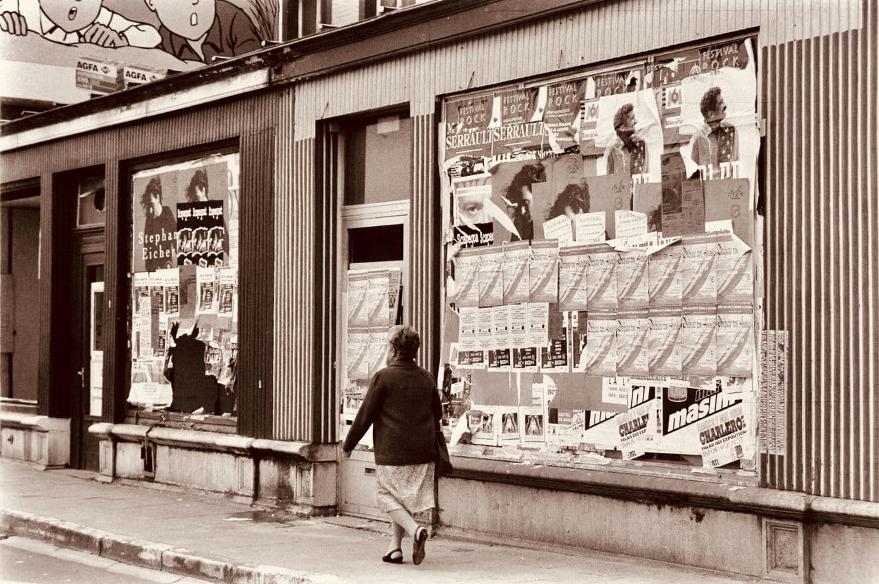 Historical street scene of a woman walking past a wall plastered with marketing posters