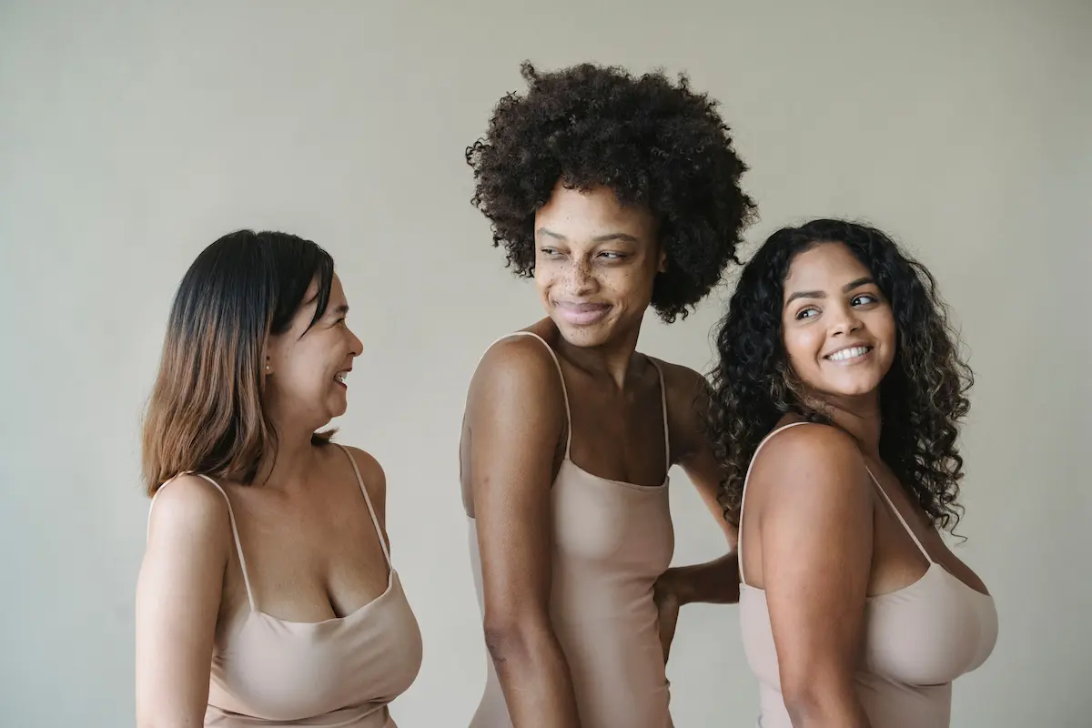 Three women of diverse backgrounds and bodytypes smiling in neutral-toned lingerie, promoting body positivity and self-confidence.